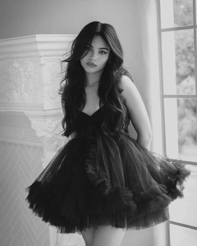 Ariana Greenblatt: Wears a short black tulle dress with ruffles, sleeveless and with a plunging neckline.…