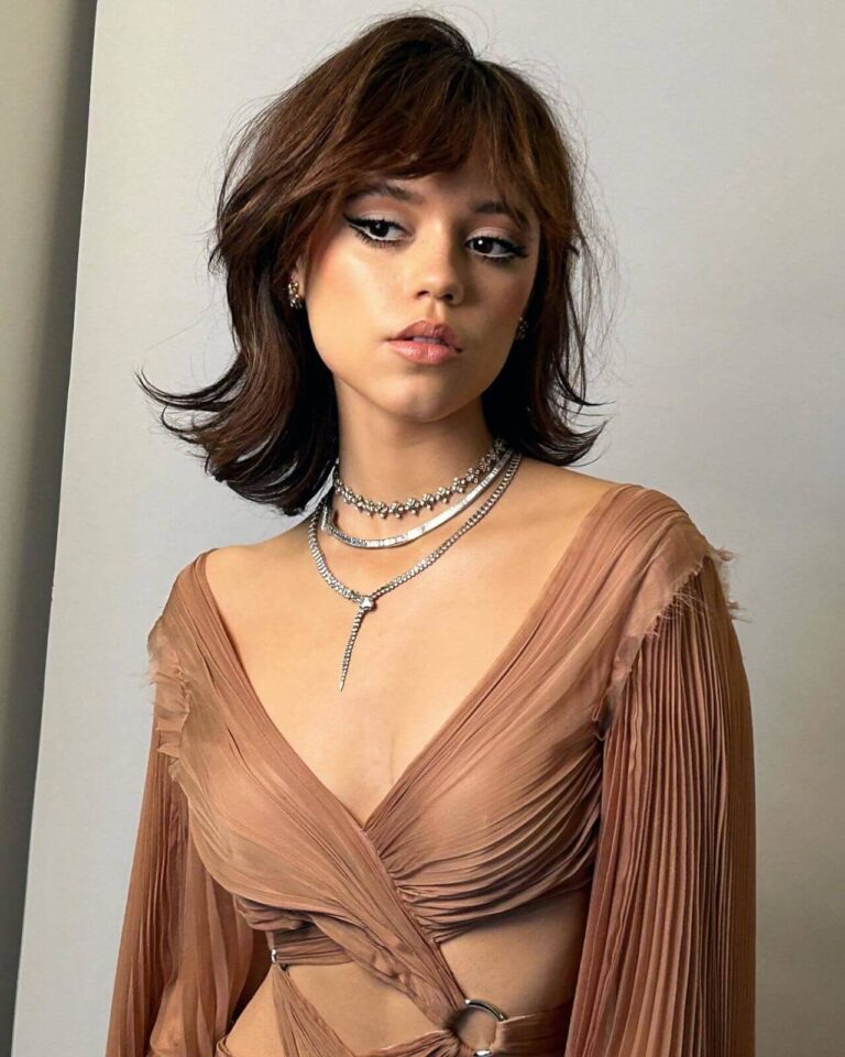 Jenna Ortega: Wears a light brown dress with a crossover design and long sleeves…