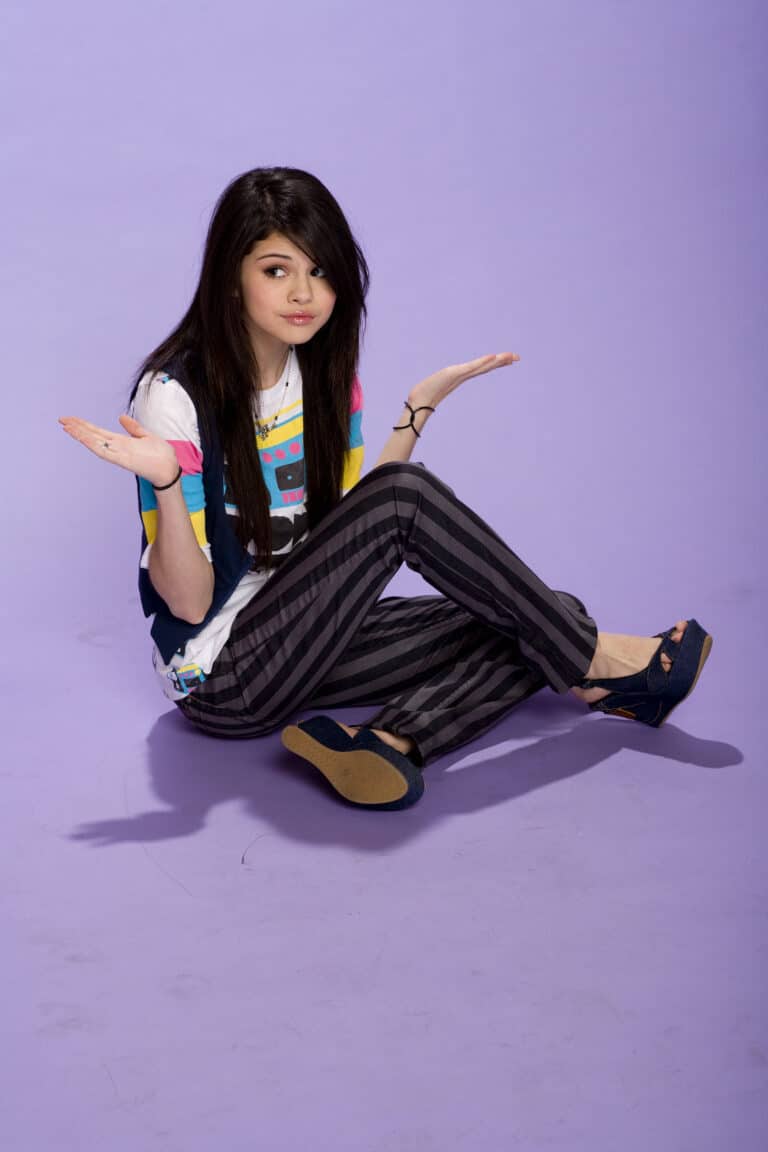 Selena Gomez: Wears a white T-shirt with colorful prints and a blue vest. The pants are…
