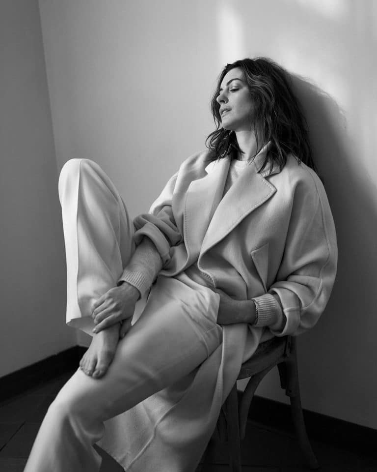 Anne Hathaway: Wearing a light-colored coat and wide pants, both in pale tones. No shoes. Her…