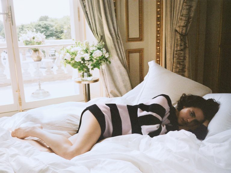 Margaret Qualley: Wears a black-and-white horizontal striped sweater, lying on a bed…