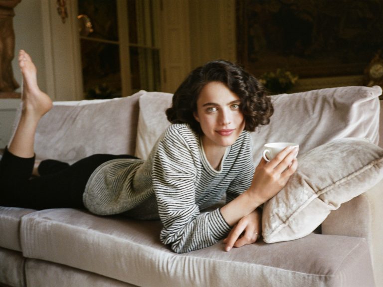 Margaret Qualley: Lying on a sofa, barefoot. Wears a white striped sweater and…