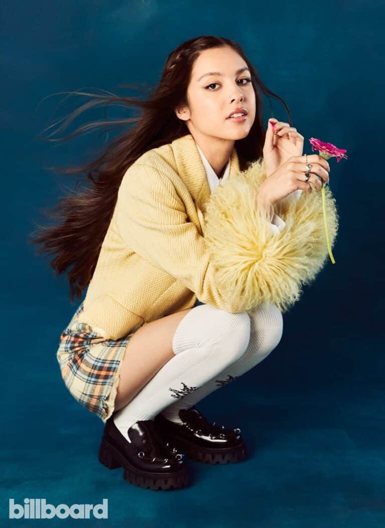 Olivia Rodrigo: Against a blue background, wearing a yellow coat with plush sleeves and a…