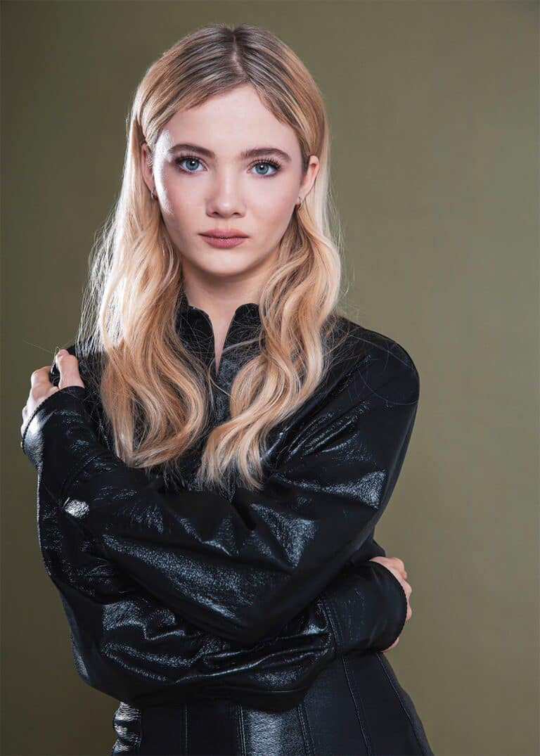Freya Allan: Long blonde hair, dressed in a shiny black jacket. Wears subtle makeup…