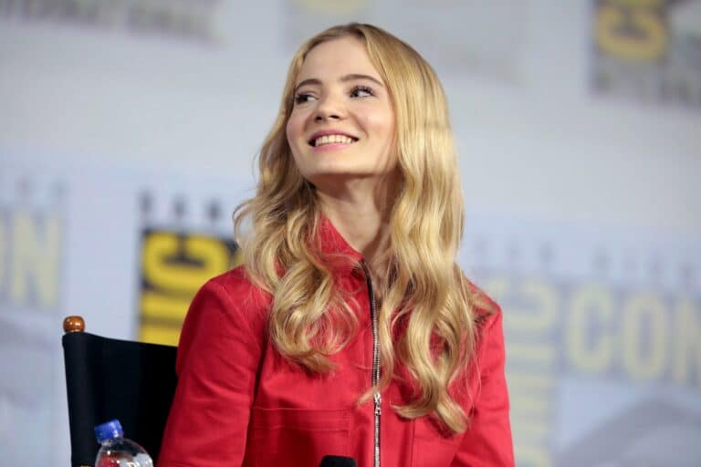Freya Allan: Wavy blonde hair, wearing a red jacket. Sitting in a chair in front…