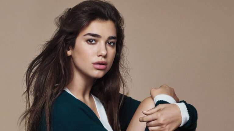 Dua Lipa: Tiene el cabello largo y suelto. Lleva un suéter azul oscuro con detalles blancos.…