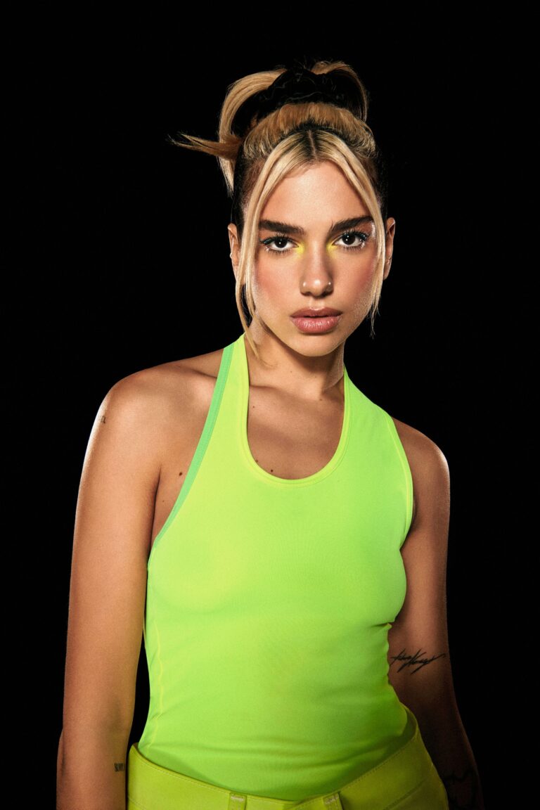 Dua Lipa: Cabello recogido con mechones delanteros sueltos, maquillaje con sombra amarilla. Lleva una camiseta verde…