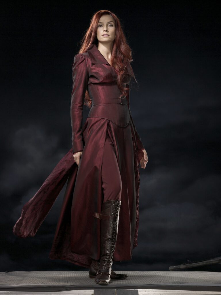 Famke Janssen: A person with long red hair wears an elegant dark red dress that…