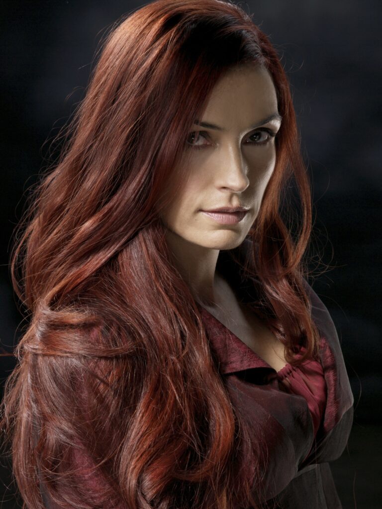 Famke Janssen: Long red hair, wearing a dark, elegant outfit. The background…