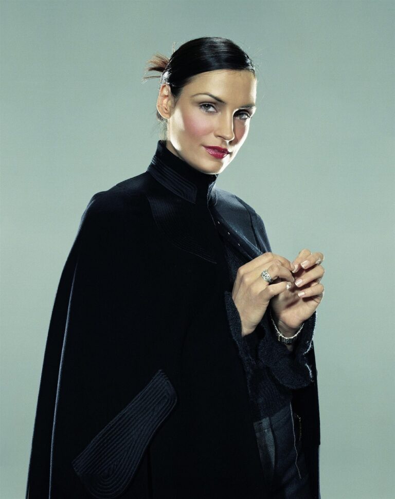 Famke Janssen: Against a neutral background, wears a dark coat over a blouse in the same shade.…