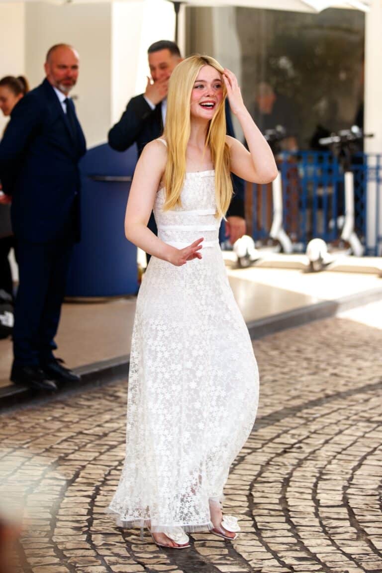 Elle Fanning: Wearing a long white lace dress, sleeveless. Her hair is blonde and…
