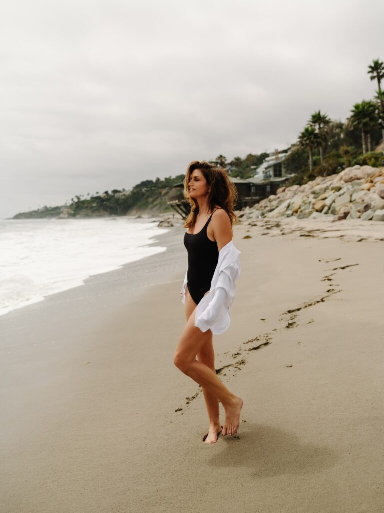 Cindy Crawford: Wearing a black swimsuit and an unbuttoned white shirt. Barefoot, walking…