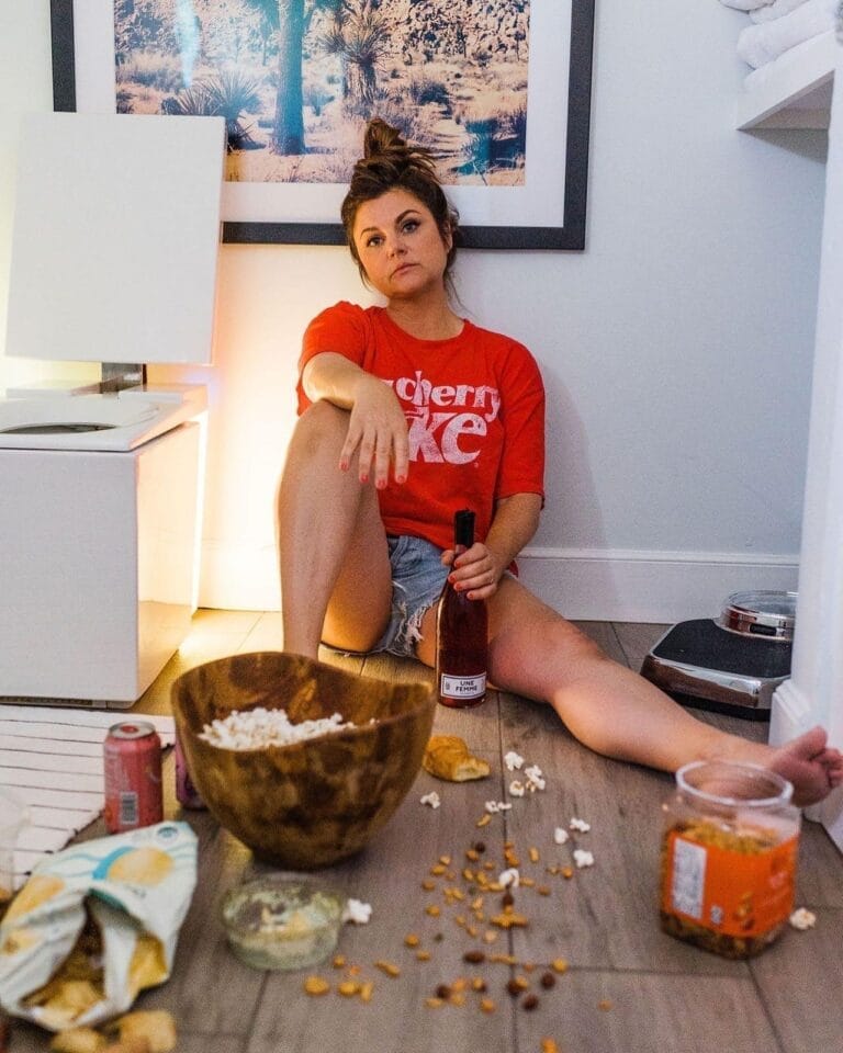Tiffani Thiessen: Lleva una camiseta roja con texto blanco y shorts de mezclilla. Está descalza, sentada…