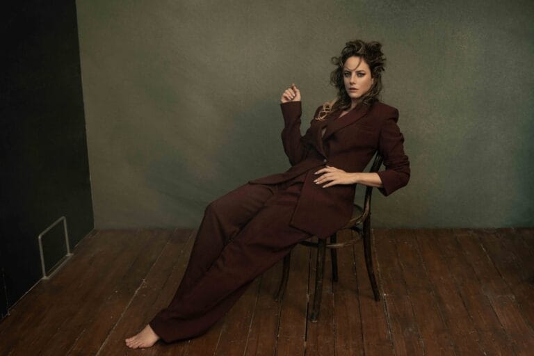 Kaya Scodelario: Wearing a dark brown tailored suit, reclining on a wooden chair on a…