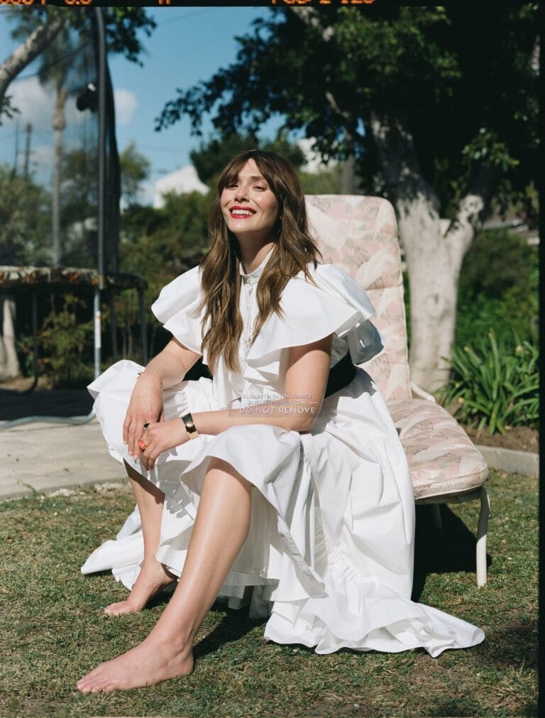 Elizabeth Olsen: Wide white dress with ruffles and short sleeves. Wears a bracelet on one wrist…