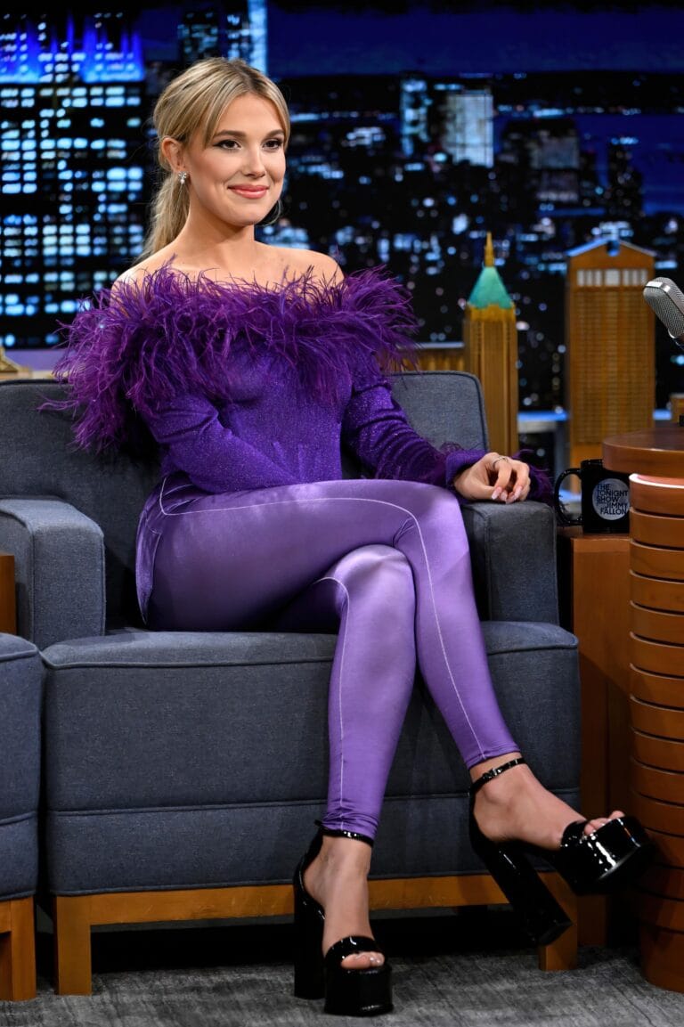 Millie Bobby Brown: Wears a purple-toned outfit made up of a shiny top with feathers on…