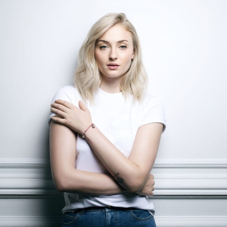 Sophie Turner: Wears a white T-shirt and blue jeans, with blonde hair worn loose. Wears…