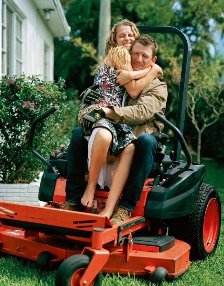 Teresa Palmer: Sits on a lawn mower in a garden. Wears a brown jacket and pants…