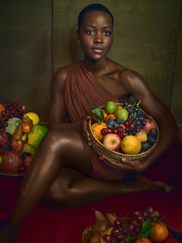 Lupita Nyong’o: Lleva una tela marrón que cubre un hombro, sosteniendo una canasta llena de frutas…