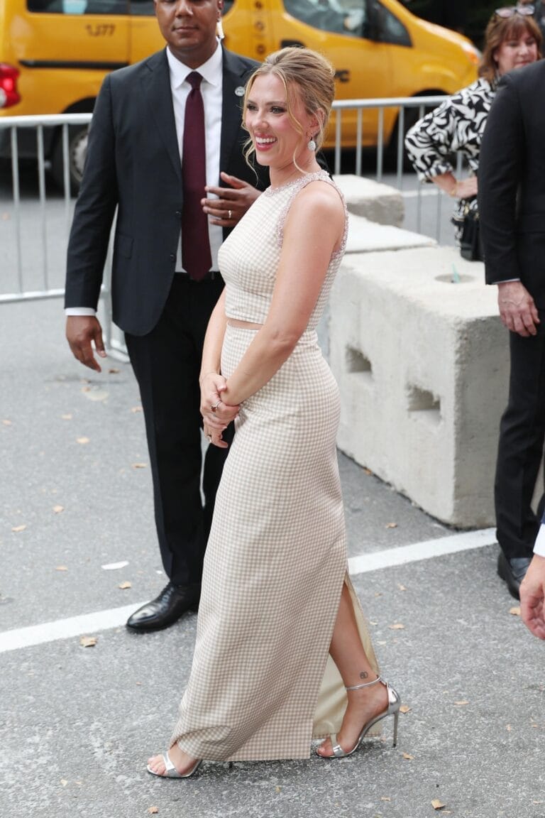 Scarlett Johansson: Wears a long plaid dress in beige and white tones, fitted at the…