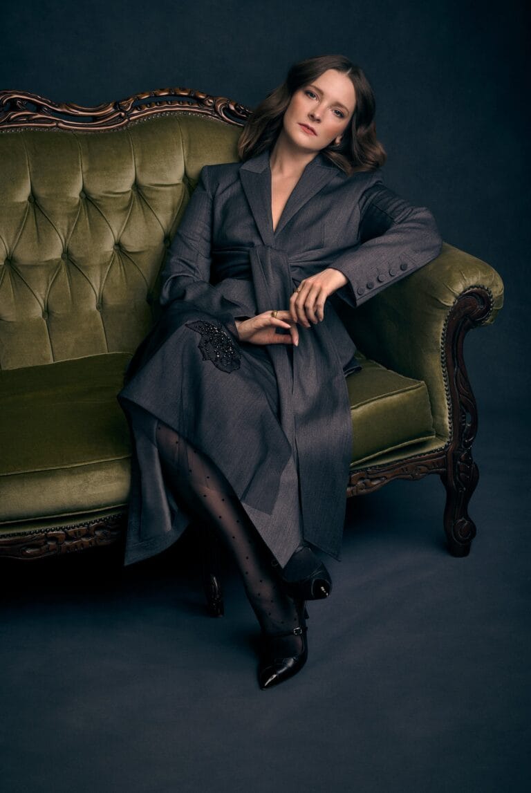 Morfydd Clark: Sentado en un sofá verde de estilo clásico, lleva un abrigo gris elegante y…