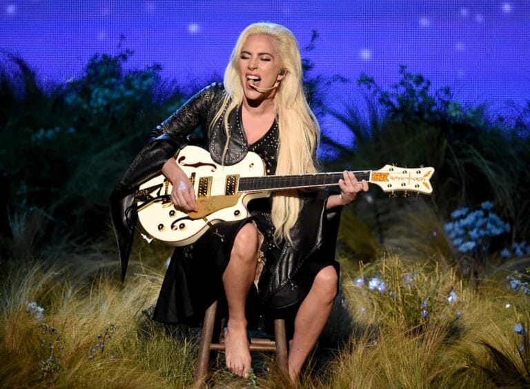 Lady Gaga: Holding a guitar, she wears a shiny black dress and a jacket...