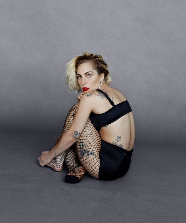 Lady Gaga: Con un fondo gris liso, viste un conjunto negro que deja al descubierto varios…