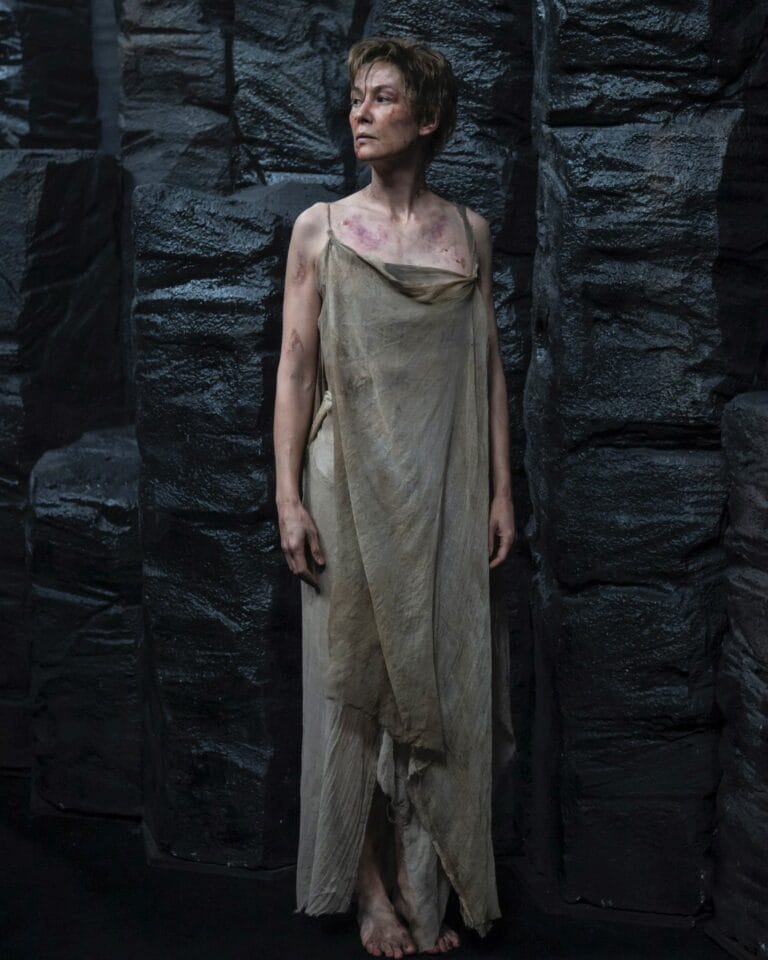 Rosamund Pike: Se encuentra en un entorno oscuro y rocoso. Lleva un vestido largo y desgastado,…