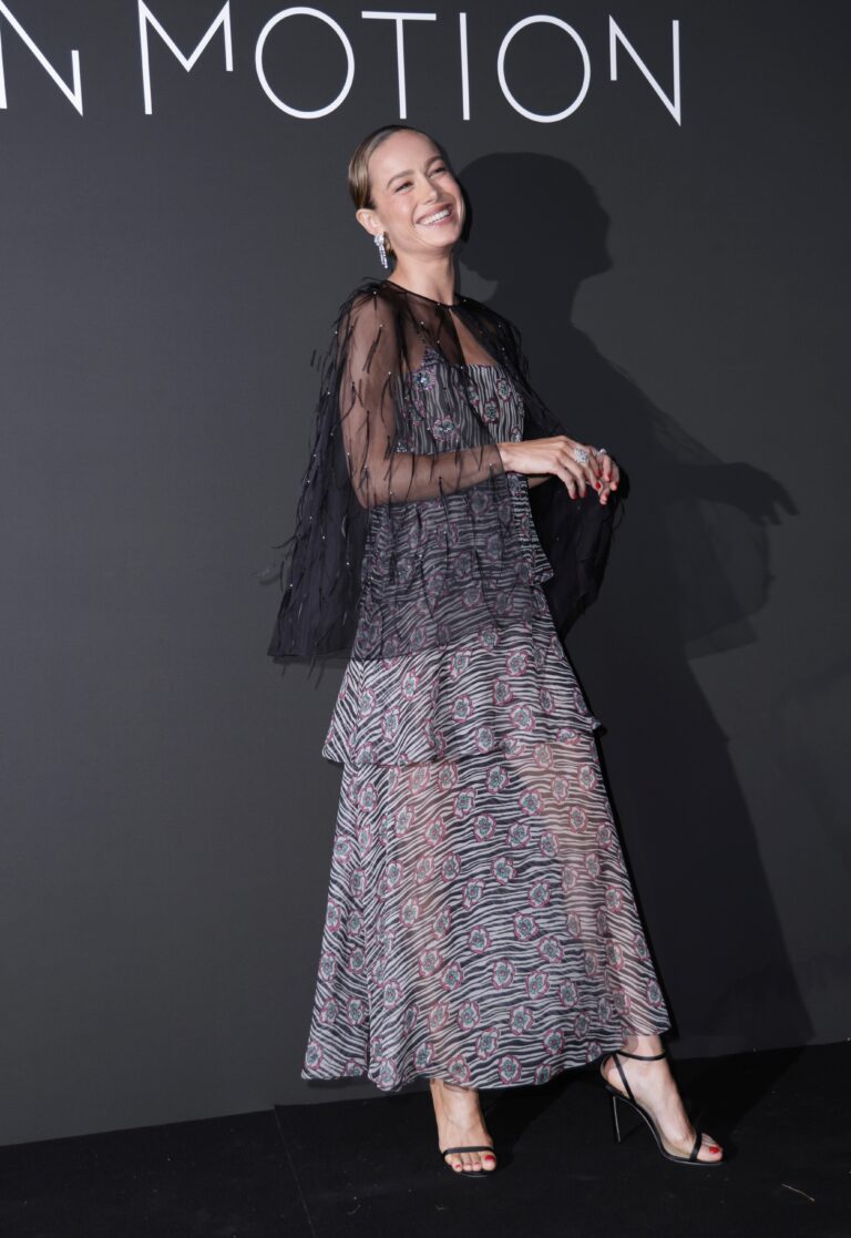 Brie Larson: Wears an elegant long dress with a floral print and sheer details. Wears sandals…