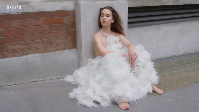 Emilia Clarke: Sitting on the ground next to a brick and concrete wall, wearing a…