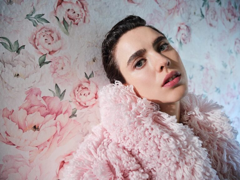 Margaret Qualley: Wears a thick, fluffy light pink coat. In the background there is a…