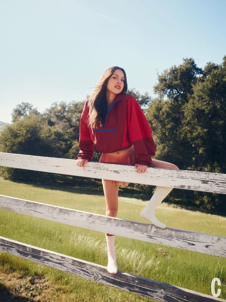 Olivia Rodrigo: Wears a red jacket with blue details and a brown miniskirt. Wears socks…