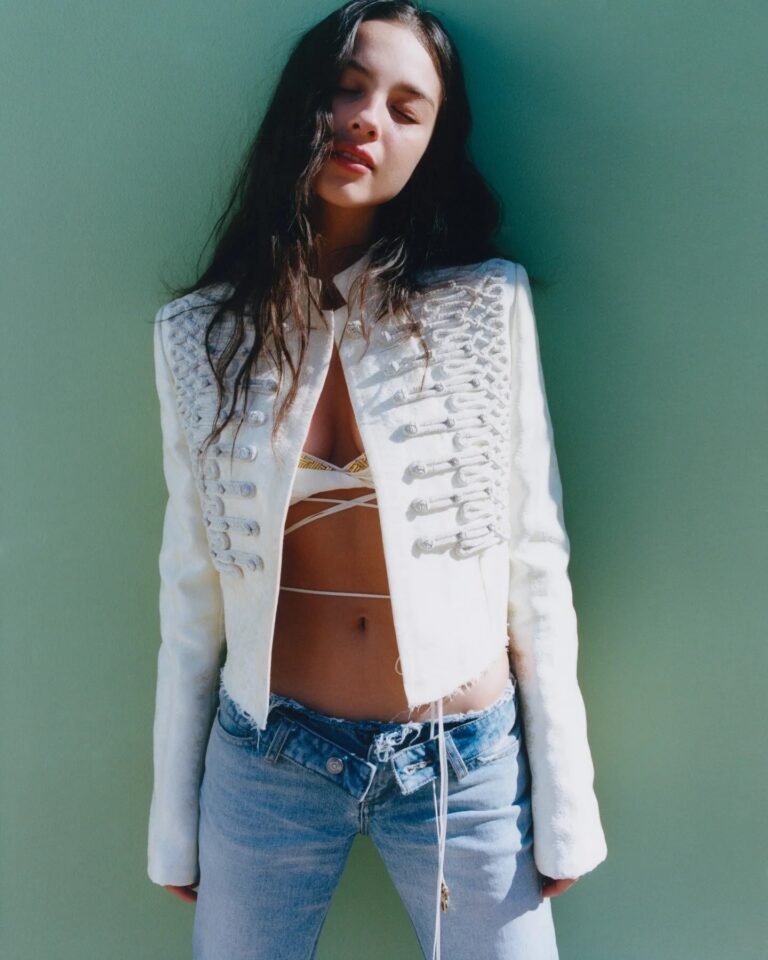 Olivia Rodrigo: Wears a white jacket with lace-up details and a strappy top underneath.…