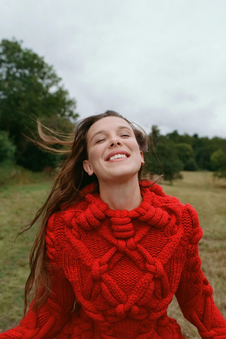 Millie Bobby Brown: Wearing a thick red knit sweater with braided details, long hair and…