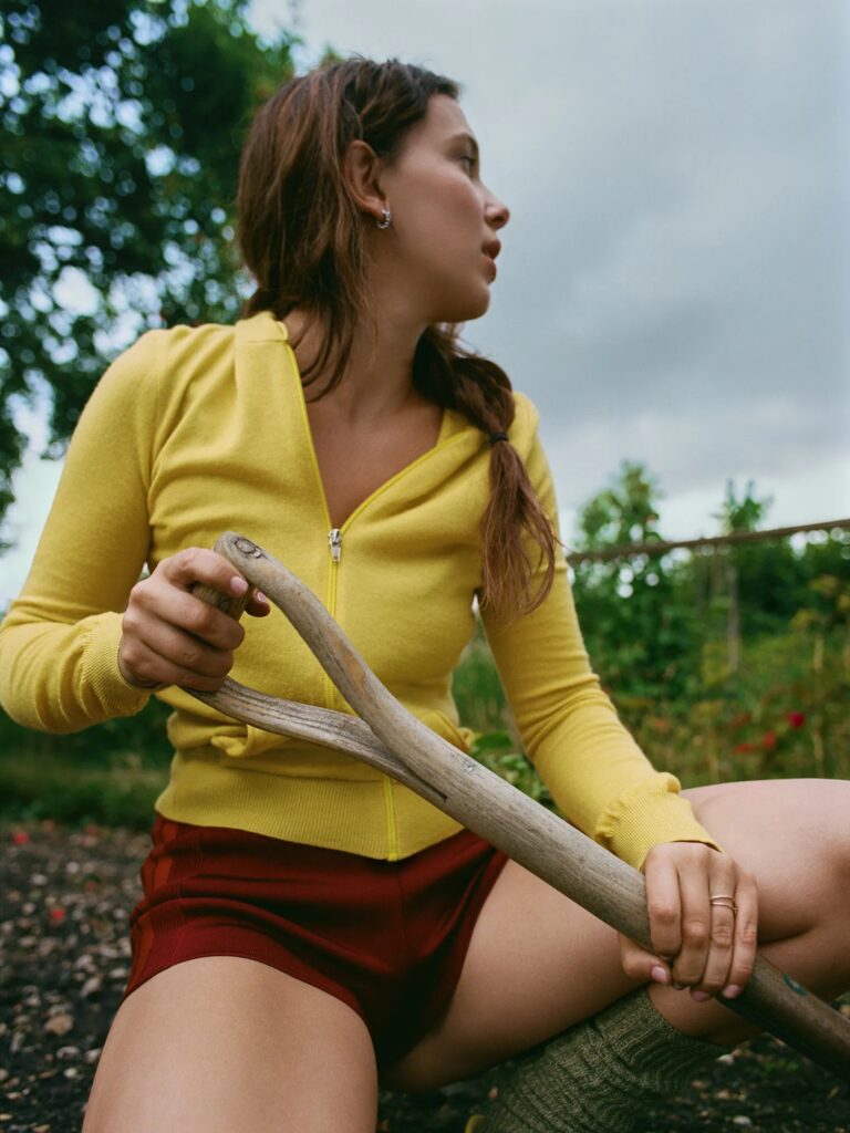 Millie Bobby Brown: Wears a yellow zip-up jacket and red shorts. Hair pulled back…