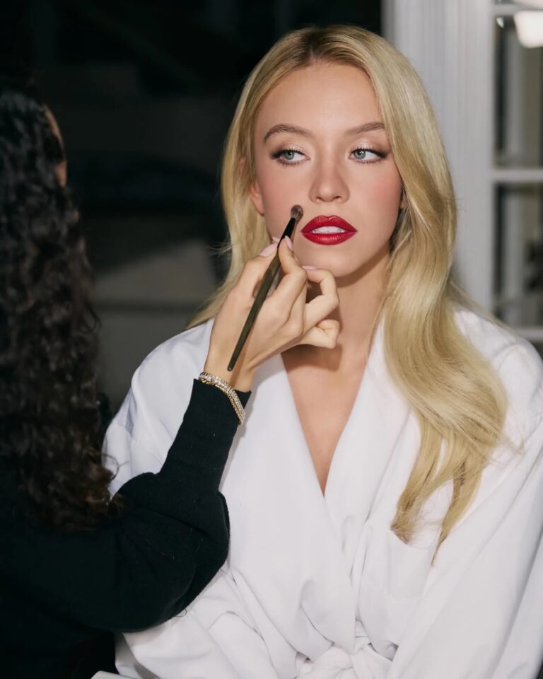 Sydney Sweeney: Está sentada tranquila mientras alguien le aplica maquillaje. Lleva una bata blanca y tiene…