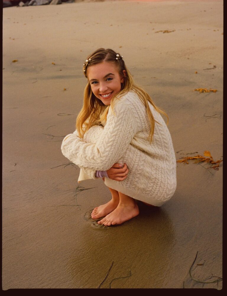 Sydney Sweeney: Está en la playa, agachada sobre la arena. Viste un suéter de punto color…