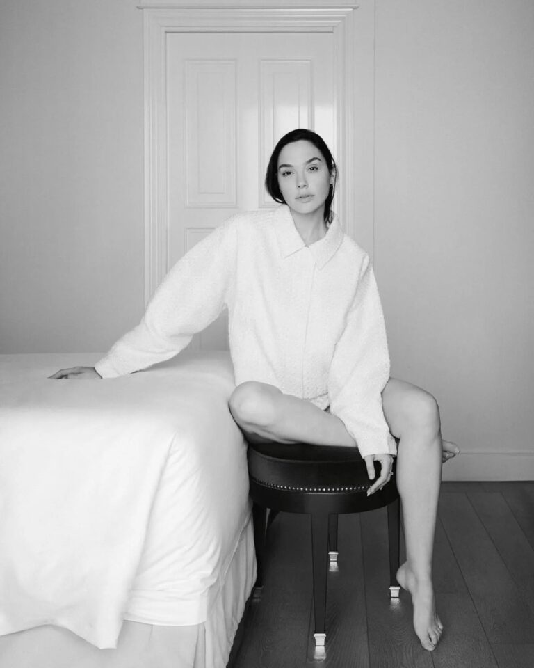 Gal Gadot: Wears a soft-textured, light-colored shirt. Is barefoot, sitting on a…