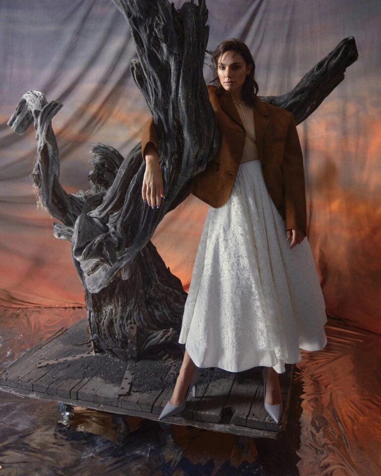 Gal Gadot: Soft light falls on a figure in a brown jacket over a long white dress…