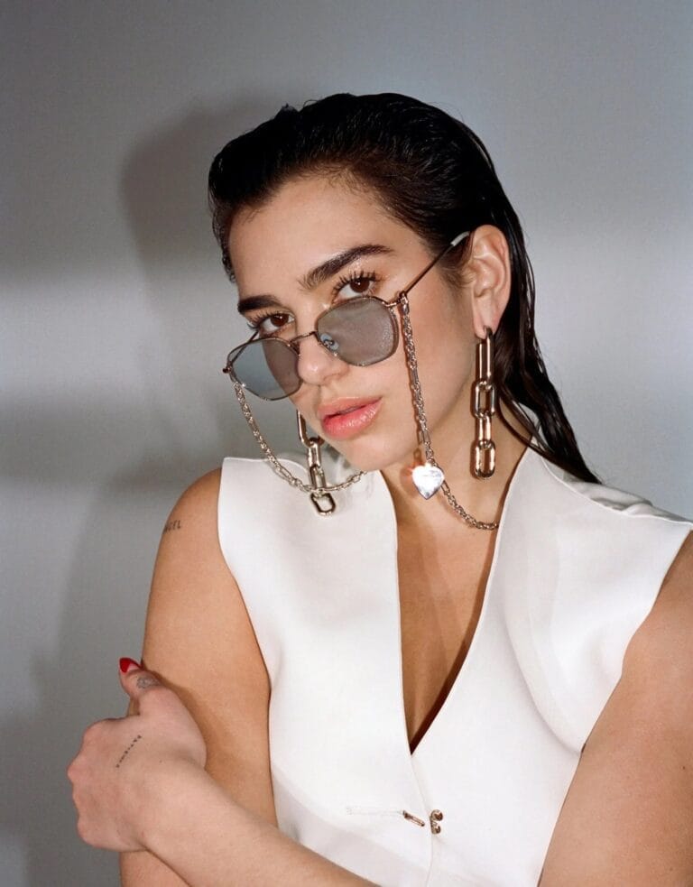 Dua Lipa: Luce una blusa blanca con un corte moderno y asimétrico, mientras se apoya levemente…