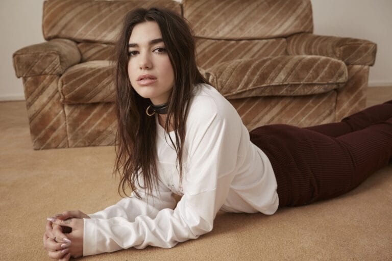 Dua Lipa: Está tumbada sobre una alfombra suave, con un estilo relajado, vistiendo una camiseta blanca…