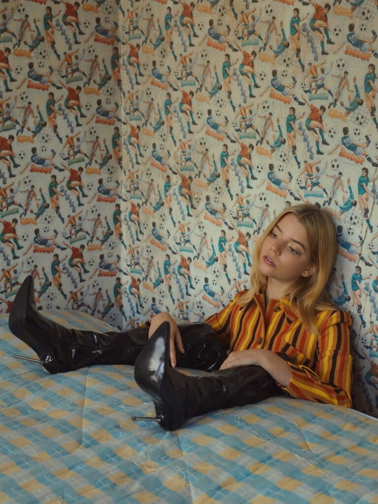 Anya Taylor-Joy: Poses seated on a bed with checkered sheets, wearing a striped blouse…