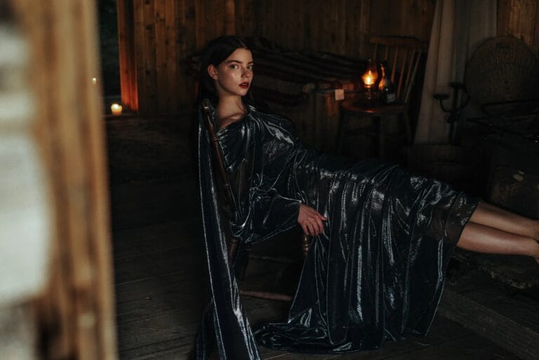 Anya Taylor-Joy: Wears a long, shiny dress in dark tones with draping that highlights…