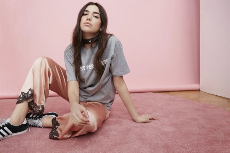 Dua Lipa: Está sentada en una alfombra rosa, con una postura relajada, las piernas flexionadas y…