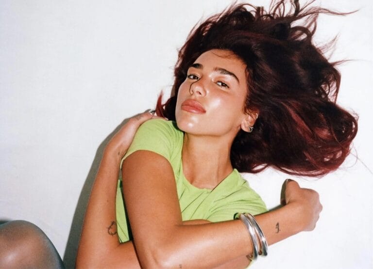 Dua Lipa: Posa tumbada sobre una superficie blanca, abrazando sus piernas mientras su cabello cae en…