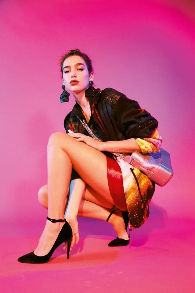 Dua Lipa: Posa en cuclillas, luciendo un vestido corto de tonos vibrantes con detalles brillantes, complementado…