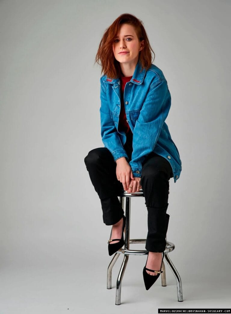 Rachel Brosnahan: She wears a blue denim jacket over a red blouse, seated on a stool…