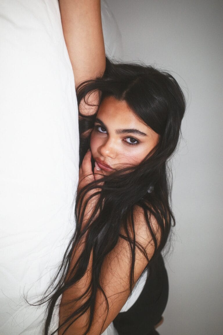 Ariana Greenblatt: Poses from the side, with her body slightly reclined on a white surface, while her…