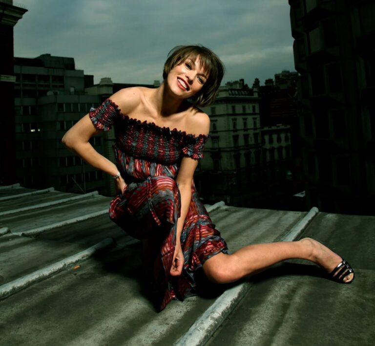 Milla Jovovich: Un vestido de color rojo y negro con estampado, de hombros descubiertos. Lleva sandalias…