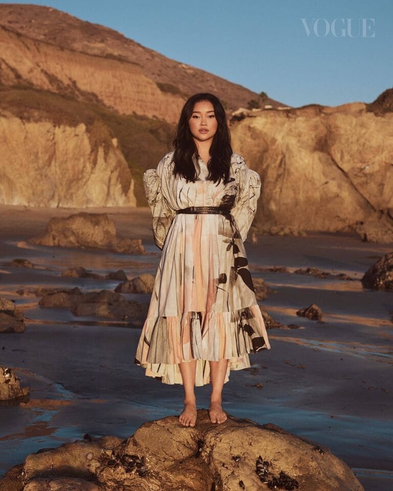Lana Condor: Poses on a rock at the beach, barefoot, in a long dress in shades of…